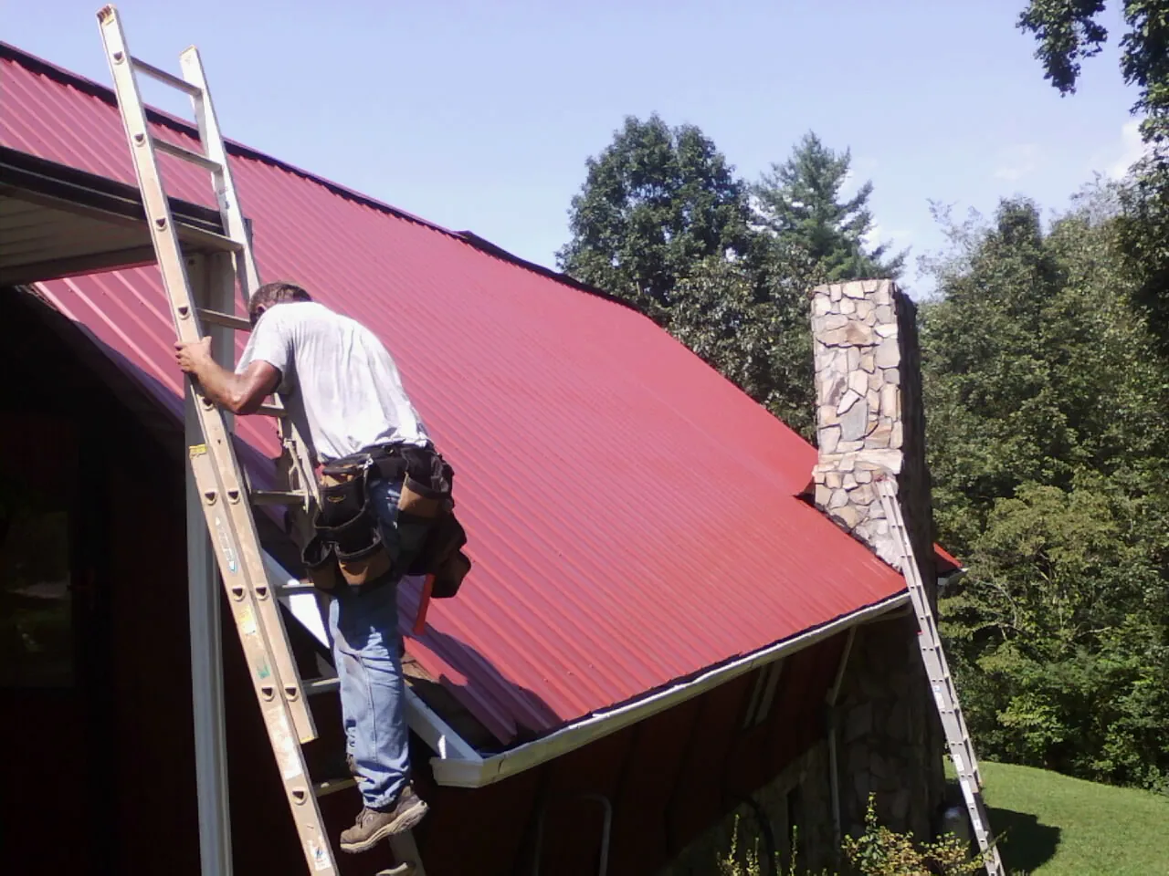 Roofer on a ladder beside a red metal roof and stone chimney — Soffit & Fascia Repair by Roofing Brothers in Paulsboro